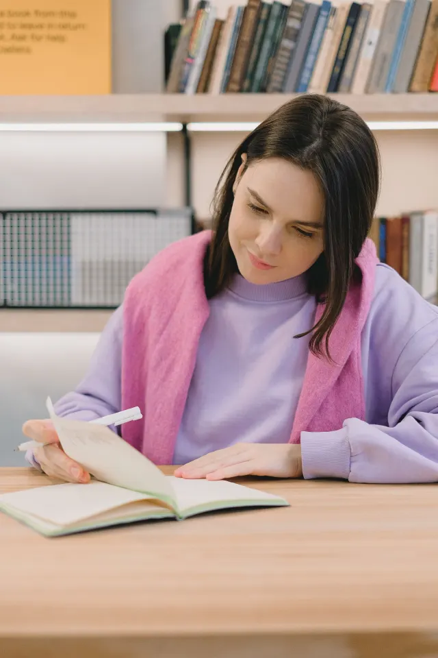A woman in a lilac jumper studying at a desk with an open notebook